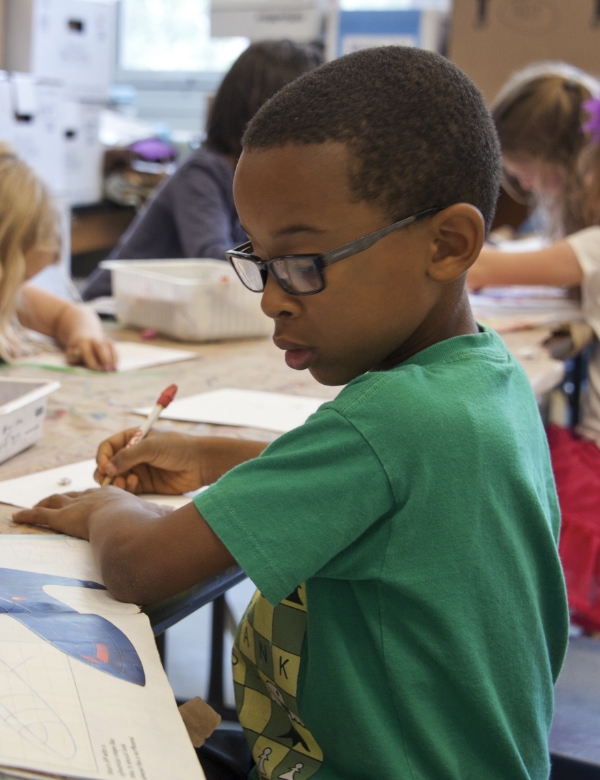 boy with glasses seated at a school desk engaged in drawing